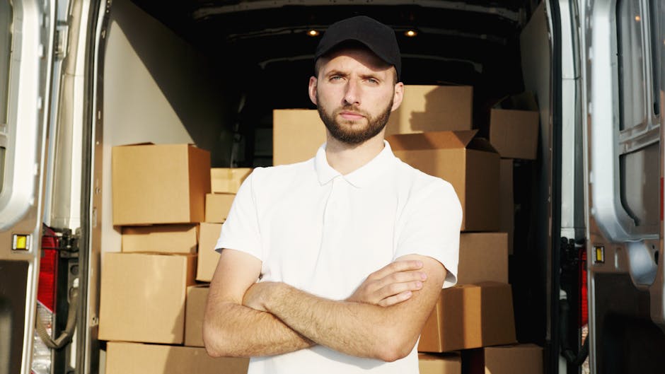 A man, dressed in a white T-shirt, jeans, and a black cap, is seen carrying a stack of three cardboard boxes outside a white commercial van parked on a residential street. The van is positioned parall