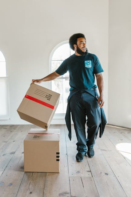 A man with dark curly hair and a beard is inside a bright, empty room with large arched windows allowing natural light to fill the space. He is dressed in a navy blue T-shirt with a logo on the chest, dark trousers, and has a jacket tied around his waist. He is actively involved in a home relocation process, holding a cardboard box with red tape in his right hand, preparing to place it onto a larger, unopened box on the wooden floor in front of him. The room has white walls and light-colored wooden flooring, providing a clean workspace for packing and moving objects. The visible packing materials and the man's posture suggest he is engaged in furniture transport and packing activities as part of a professional removals service, such as those offered by Man and Van Fulham, supporting efficient and organized house removals in Fulham.