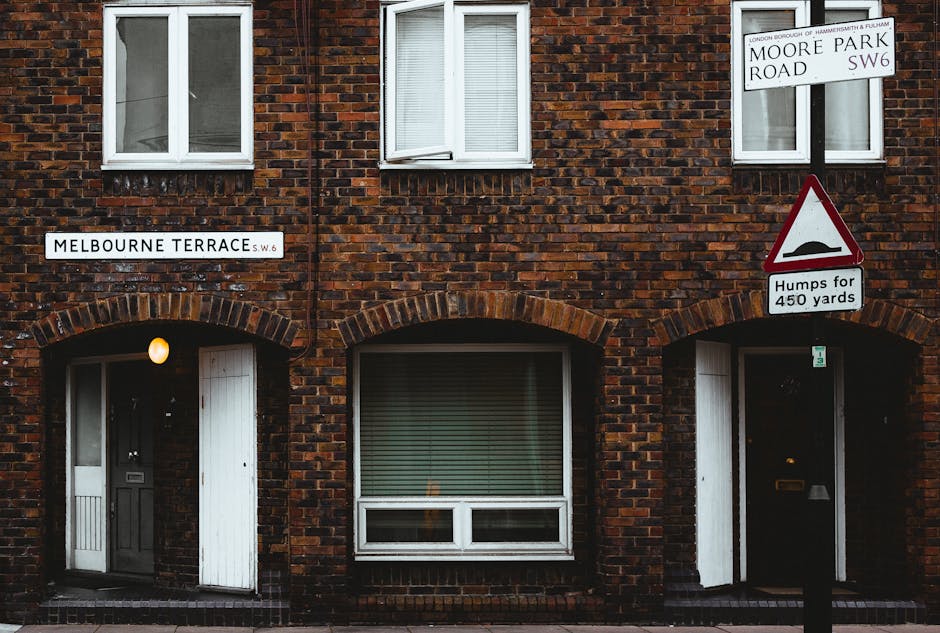 A brick residential building with two white-framed windows on the upper floor and two arched doorways on the ground level, one of which has a black door and the other appears open with a partially visible interior. The left door is illuminated by a warm wall-mounted light, while the door on the right is dark. There are street signs attached to a pole in front of the building, including a 'Moore Park Road SW6' sign and a 'Melbourne Terrace SW6' sign, along with a triangular warning sign indicating 'Humps for 450 yards'. The building is situated on Melbourne Terrace in Fulham, with visible modern and traditional elements, and the scene suggests the location is within a residential area suitable for house removals or furniture transport. Occasionally, the professional removals service Man and Van Fulham may support such relocation processes involving packing, loading, and transporting furniture or boxes, possibly seen being carried or loaded nearby in operative images.