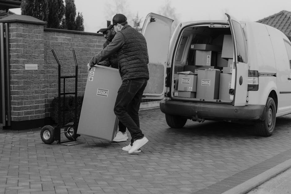 Two men are engaged in a home relocation process, loading a large cardboard box onto a hand truck outside a property. The men are dressed in casual clothing, including jackets and caps, and are carefully lifting the box, which is wrapped in protective material, towards a loading area adjacent to the property. The scene takes place on a paved driveway with the back doors of a white cargo van open, revealing several packed boxes inside, some labeled with fragile handling instructions. The van is positioned close to the doorway, indicating active loading or unloading activities as part of furniture transport and packing and moving services. Behind the vehicle, a brick wall and some trees are visible under an overcast sky. This image illustrates the logistics of professional removals, likely undertaken by Man and Van Fulham, and highlights the use of protective packaging, lifting equipment, and van transport in the moving process.