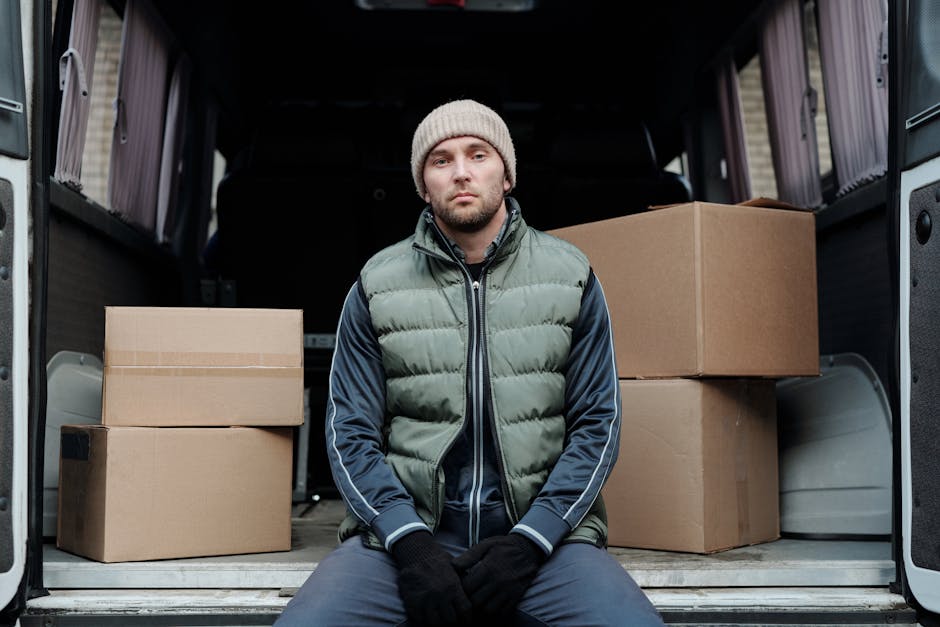 A man with a serious expression, wearing a beige knit hat, a green padded vest over a dark navy jacket, and black gloves, is seated on the edge of an open moving van. Inside the van, there are several packed cardboard boxes of various sizes, some with tape sealing the edges, and a few wrapped in protective plastic or fabric for transport. The boxes are arranged on the van's floor, with some placed directly on the metal surface and others stacked on top of each other. The van's interior is visible behind the man, showing a dark, spacious cargo area with high walls. The scene captures the loading process during a house removal or furniture transport, highlighting the careful packing and positioning of household items within the vehicle. Man and Van Fulham, a removals company, appears to facilitate the loading and unloading for home relocations, as evidenced by the context of the scene.