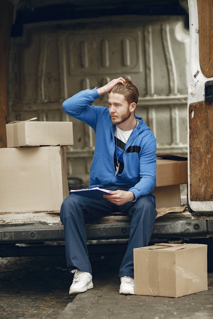 Two men are engaged in a home relocation process, loading a large cardboard box onto a hand truck outside a property. The men are dressed in casual clothing, including jackets and caps, and are carefully lifting the box, which is wrapped in protective material, towards a loading area adjacent to the property. The scene takes place on a paved driveway with the back doors of a white cargo van open, revealing several packed boxes inside, some labeled with fragile handling instructions. The van is positioned close to the doorway, indicating active loading or unloading activities as part of furniture transport and packing and moving services. Behind the vehicle, a brick wall and some trees are visible under an overcast sky. This image illustrates the logistics of professional removals, likely undertaken by Man and Van Fulham, and highlights the use of protective packaging, lifting equipment, and van transport in the moving process.